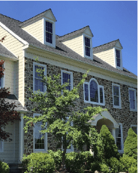 Stone house with cream siding, arched window, and dormer windows surrounded by trees under a blue sky