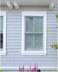 White-framed window on a light gray shingled house exterior with purple flowers below.