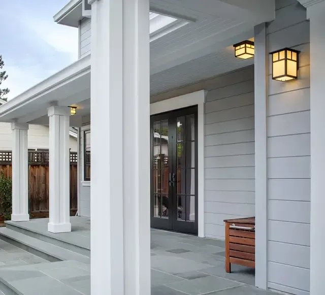 Covered front porch with white columns, gray siding, lantern lights, and a small wooden bench.