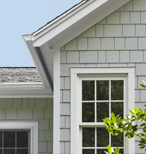 Gray-shingled house exterior with white trim, roof eaves, and windows beside green foliage