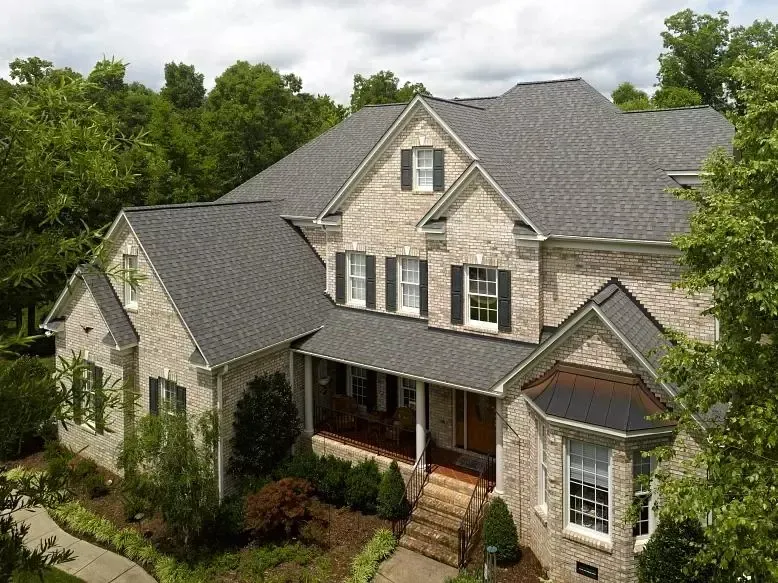 Large stone suburban house with gray roof and front porch, surrounded by trees and landscaping