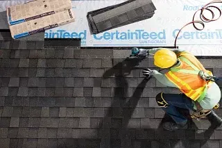 Worker in a yellow hard hat installing dark shingles on a roof with a nail gun.
