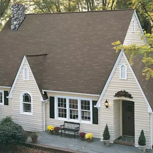 White cottage with steep brown roof, black shutters, and a front porch with flowers and bench.