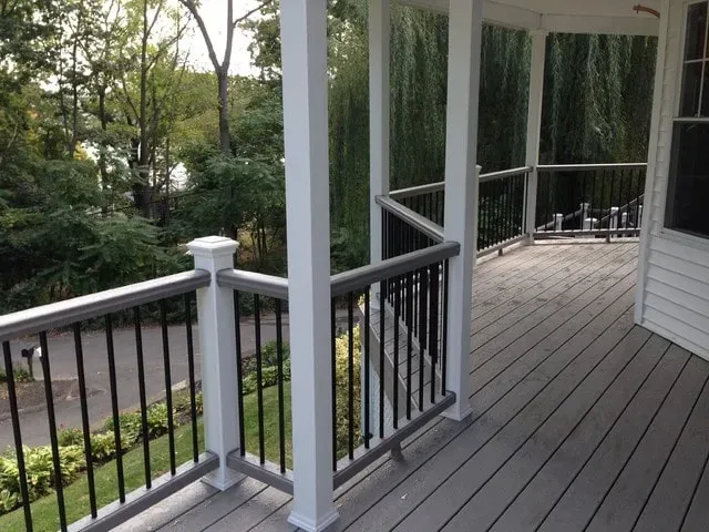 Covered porch with gray deck, white columns, and black railing overlooking a wooded yard