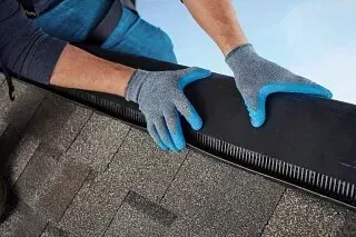 Gloved hands installing a black roof vent or edge trim on gray shingles