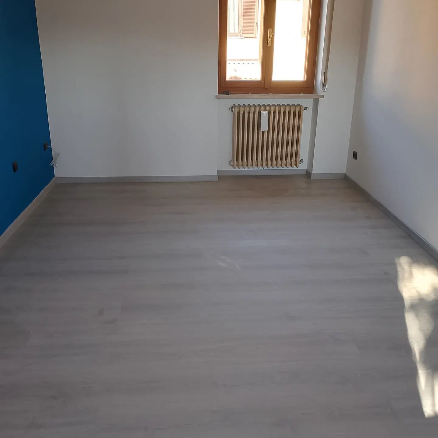 Una stanza vuota con pavimento effetto legno chiaro, pareti bianche, una parete d'accento blu scuro, una finestra e un termosifone a parete.
