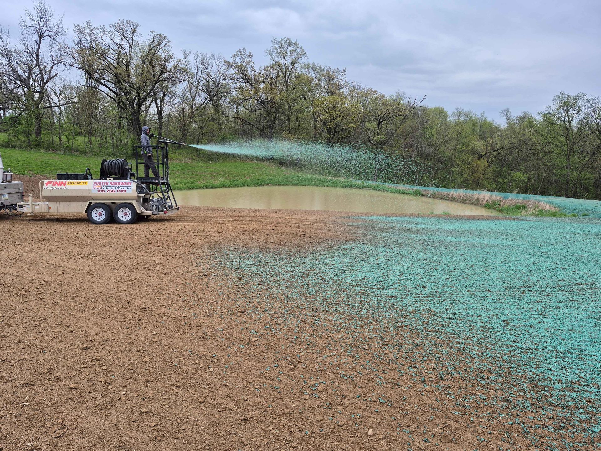 Sprayer truck spreading green liquid across a brown field near trees under a cloudy sky
