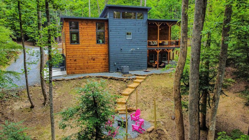 Modern home nestled in a forest with wooden stairs leading to a patio with pink chairs.
