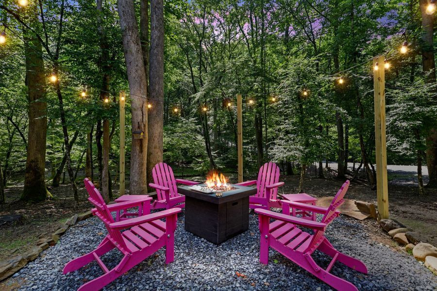 Pink chairs circle a fire pit in a wooded area, strung lights overhead.