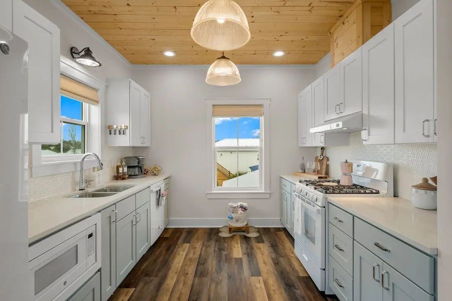 Bright white kitchen with light blue cabinets, wood ceiling and floor, two windows.