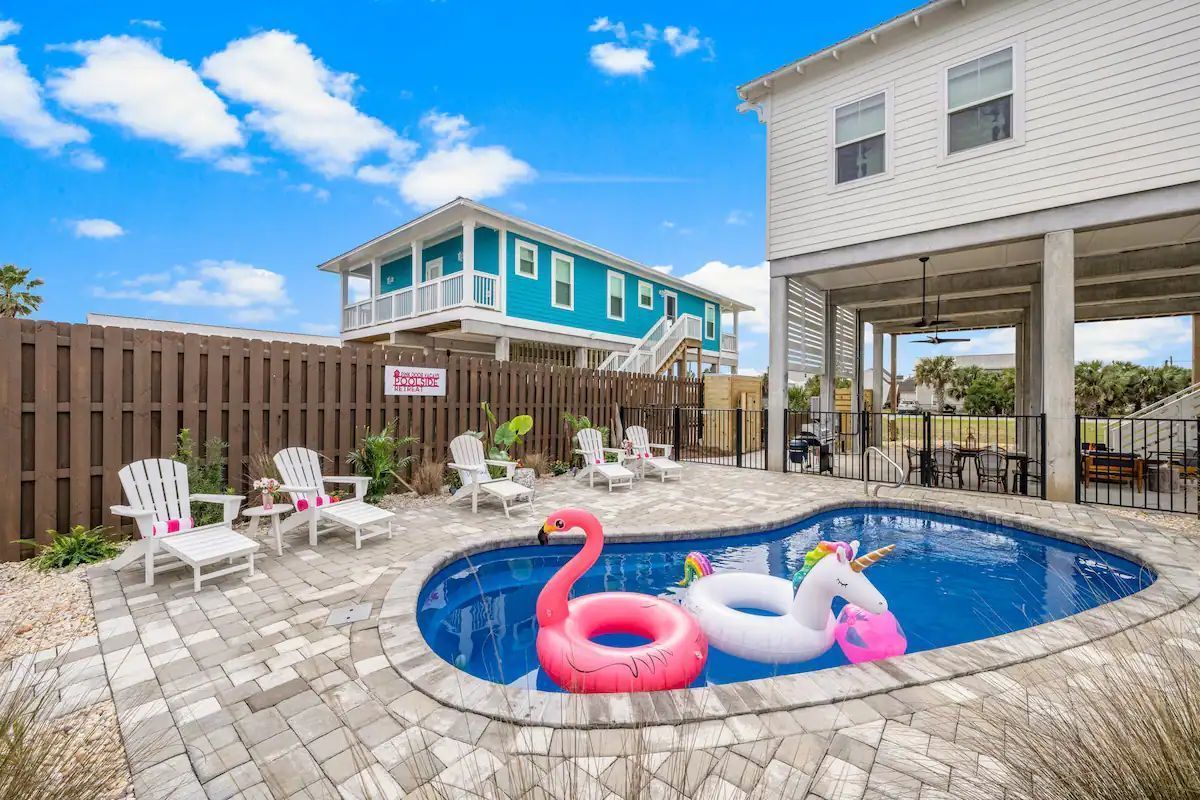 Backyard pool with inflatable flamingo and unicorn, lounge chairs, and houses. Bright blue sky.