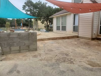 Patio with stone pavers, concrete block wall, and shade sails. House with windows visible in background.