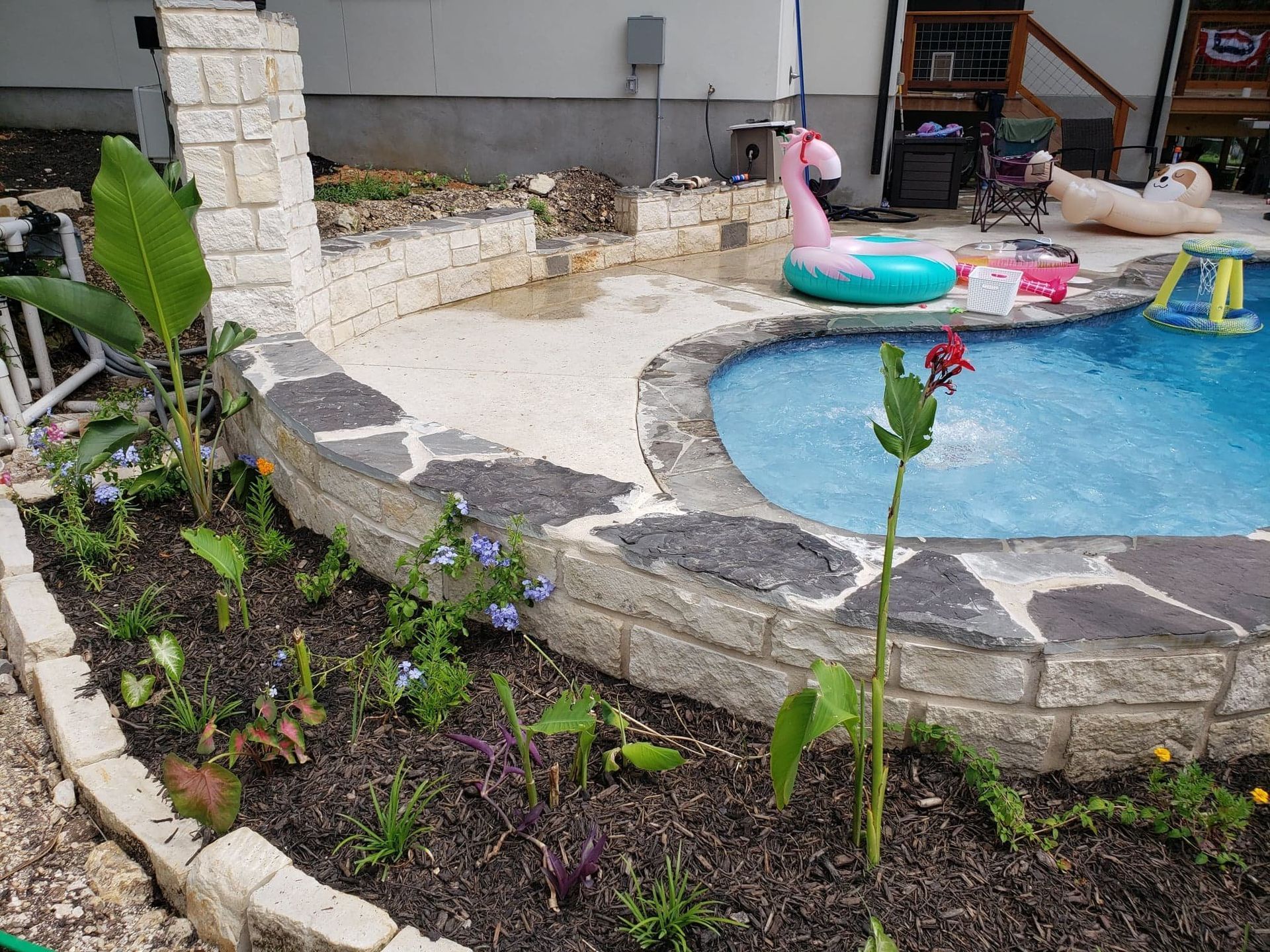 Poolside garden with stone edging and various plants; pink flamingo float in pool.