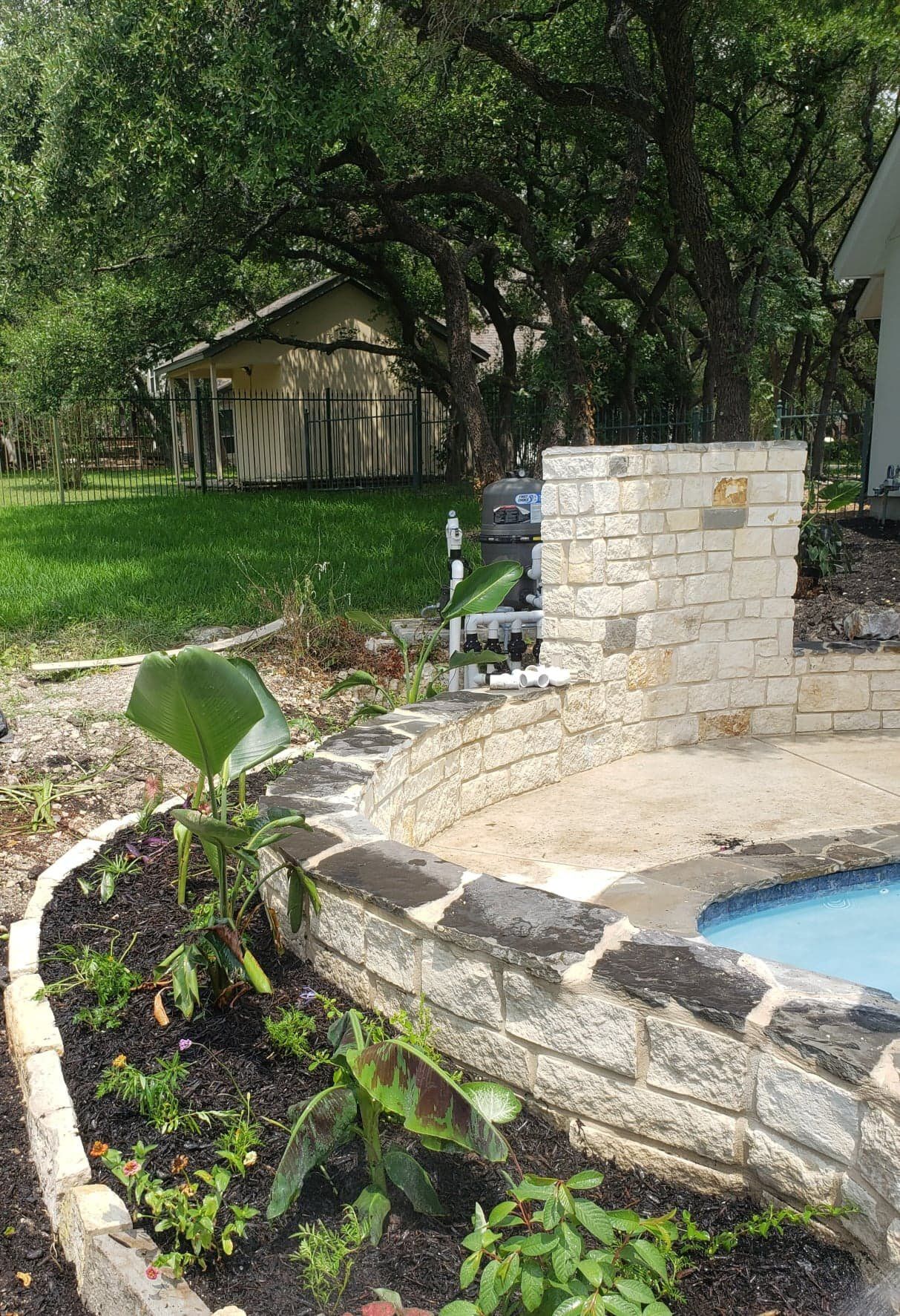 Poolside garden bed with stone walls and a building in the background, under tree cover.