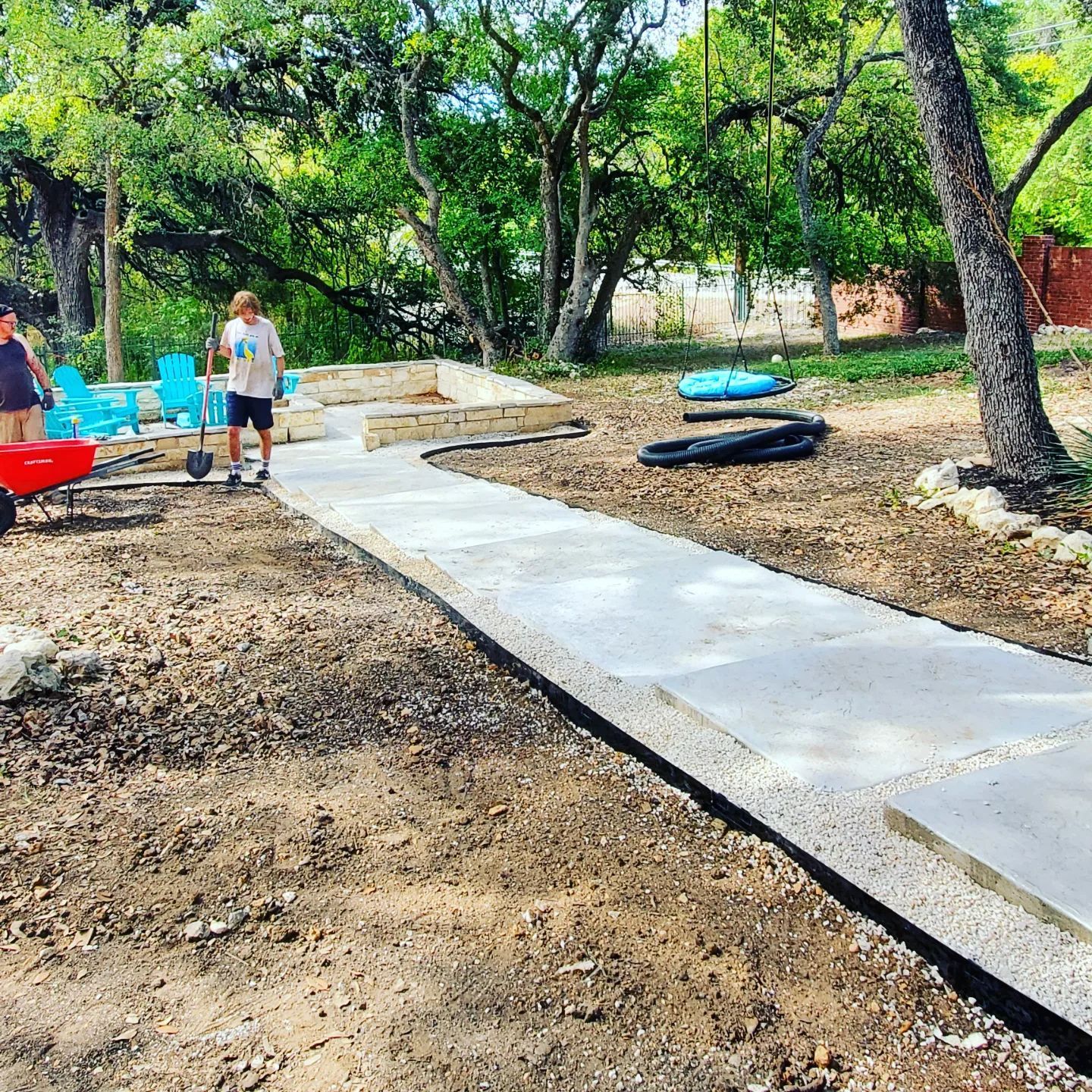 Two men working on a concrete path in a yard, trees and chairs in background.