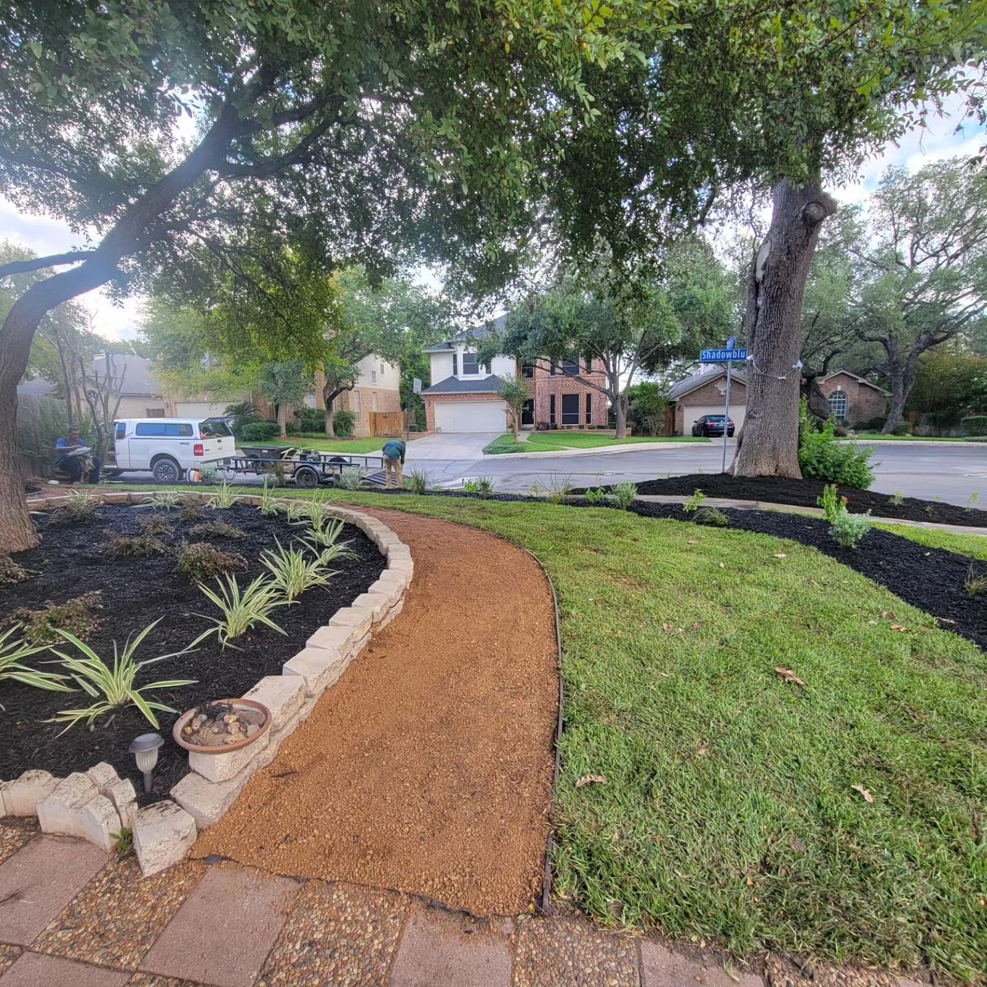 A gravel pathway curves through a front yard with mulch beds, grass, and trees. Houses are visible in the background.