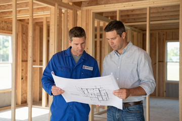 Two men reviewing a house blueprint inside an unfinished wooden frame house.