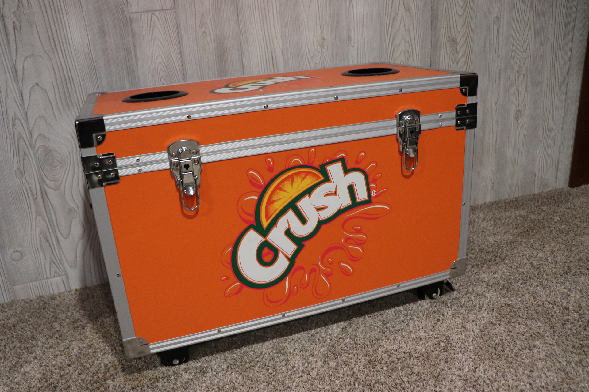 An orange crush cooler is sitting on a carpeted floor.
