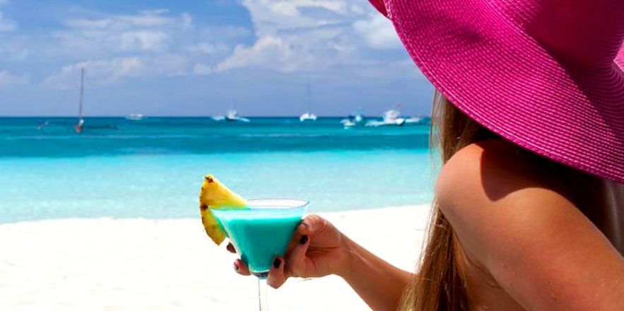 Una mujer con un sombrero rosa sostiene un cóctel azul en la playa.