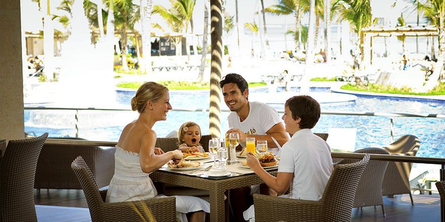 Una familia está sentada en una mesa frente a una piscina comiendo comida.