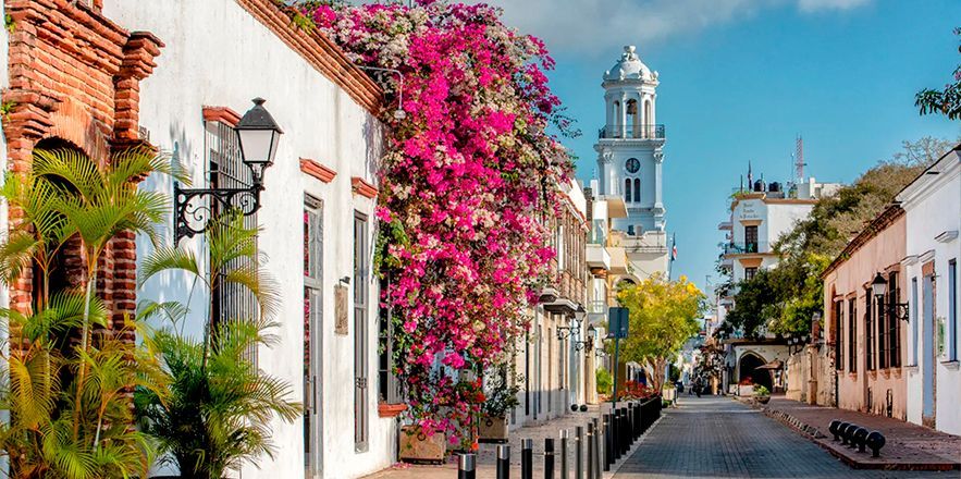 Una calle estrecha bordeada de edificios blancos y flores rosadas con una iglesia al fondo.
