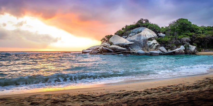 Una playa con una gran roca en medio del océano al atardecer.