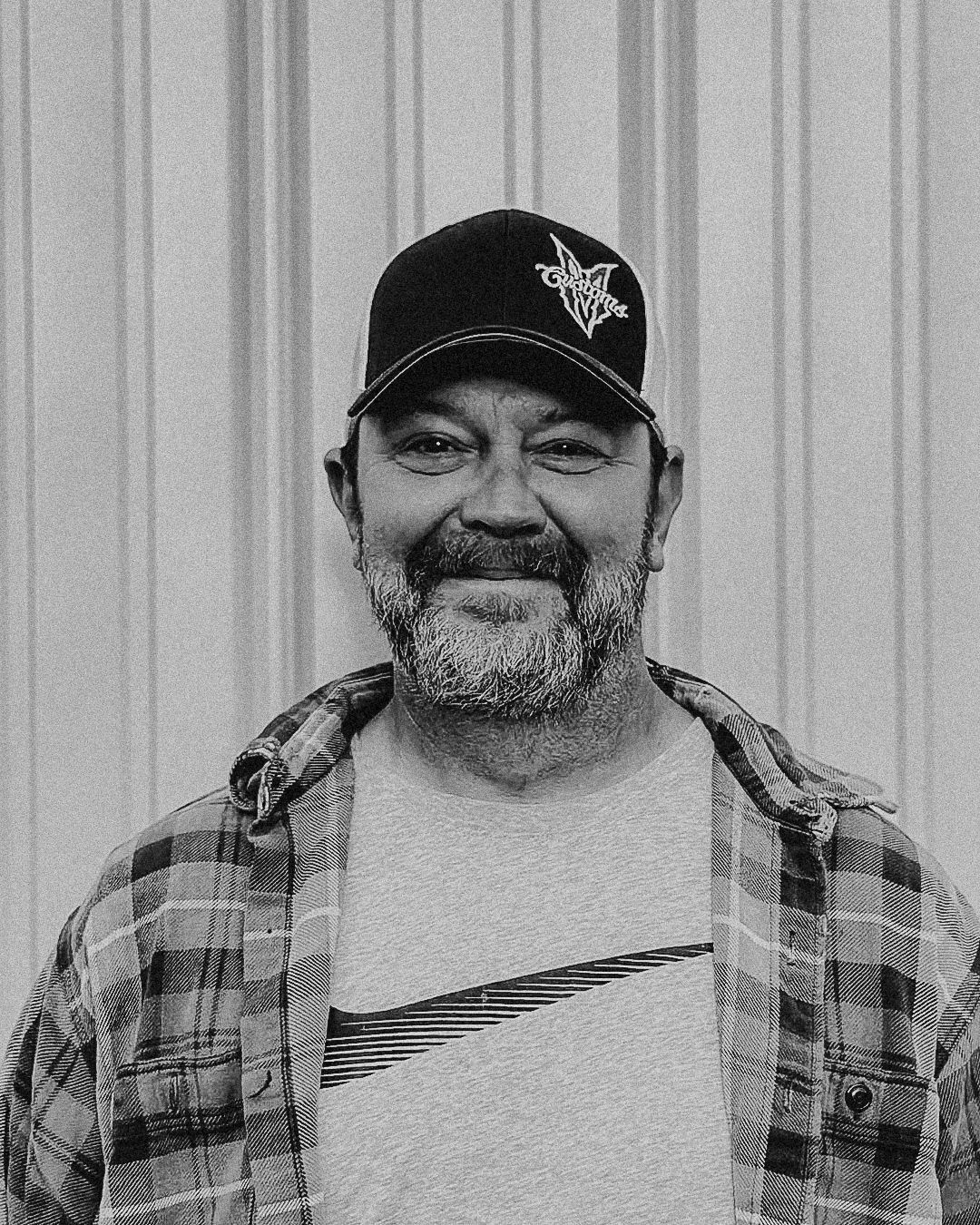 Man wearing a cap, plaid shirt, and grey t-shirt, standing in front of a corrugated metal wall.
