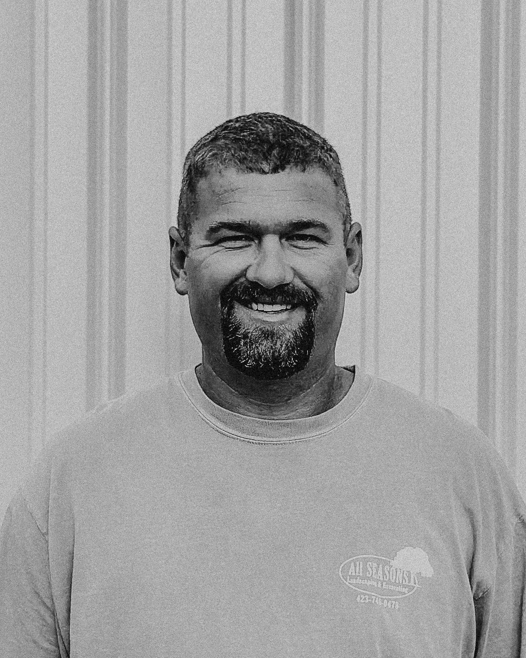 Man with short dark hair and beard smiling, wearing a gray t-shirt, standing in front of a beige wall.