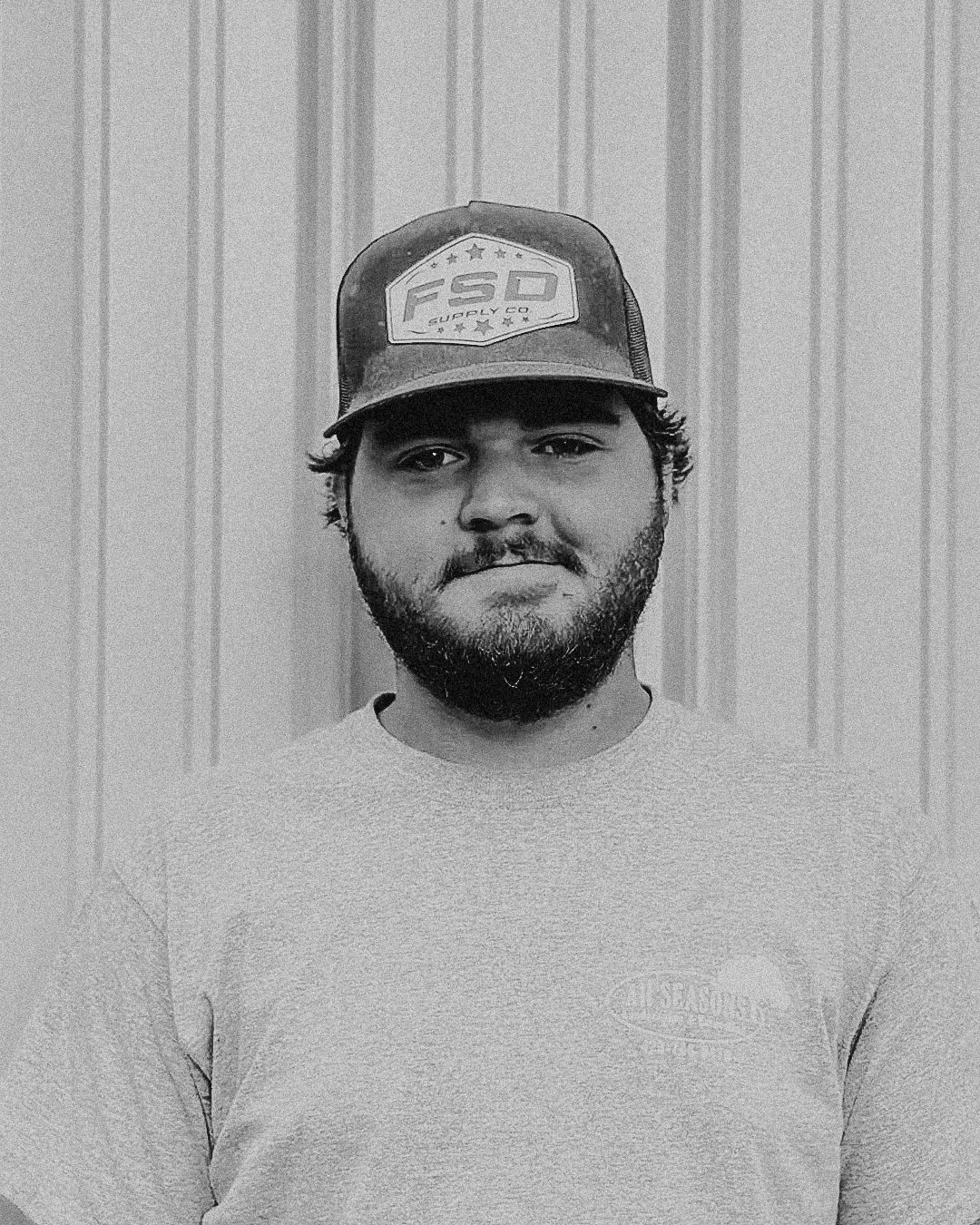 Young man with a beard wearing a hat and gray t-shirt stands in front of a corrugated metal wall.