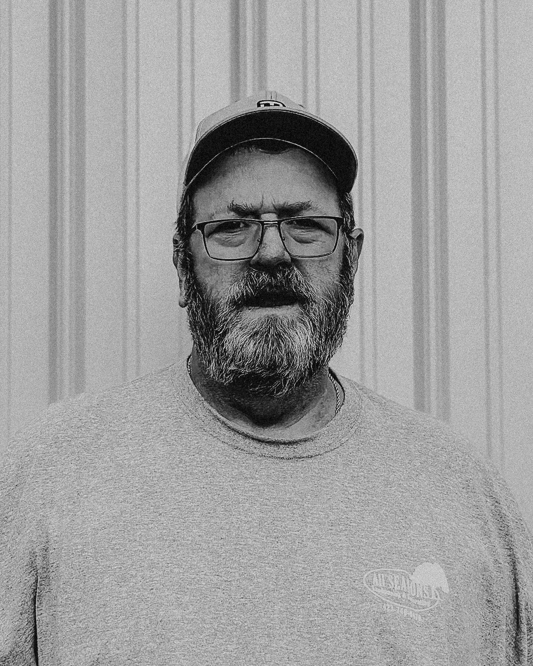 Man with beard and glasses in a gray t-shirt and cap, standing in front of a corrugated metal wall.