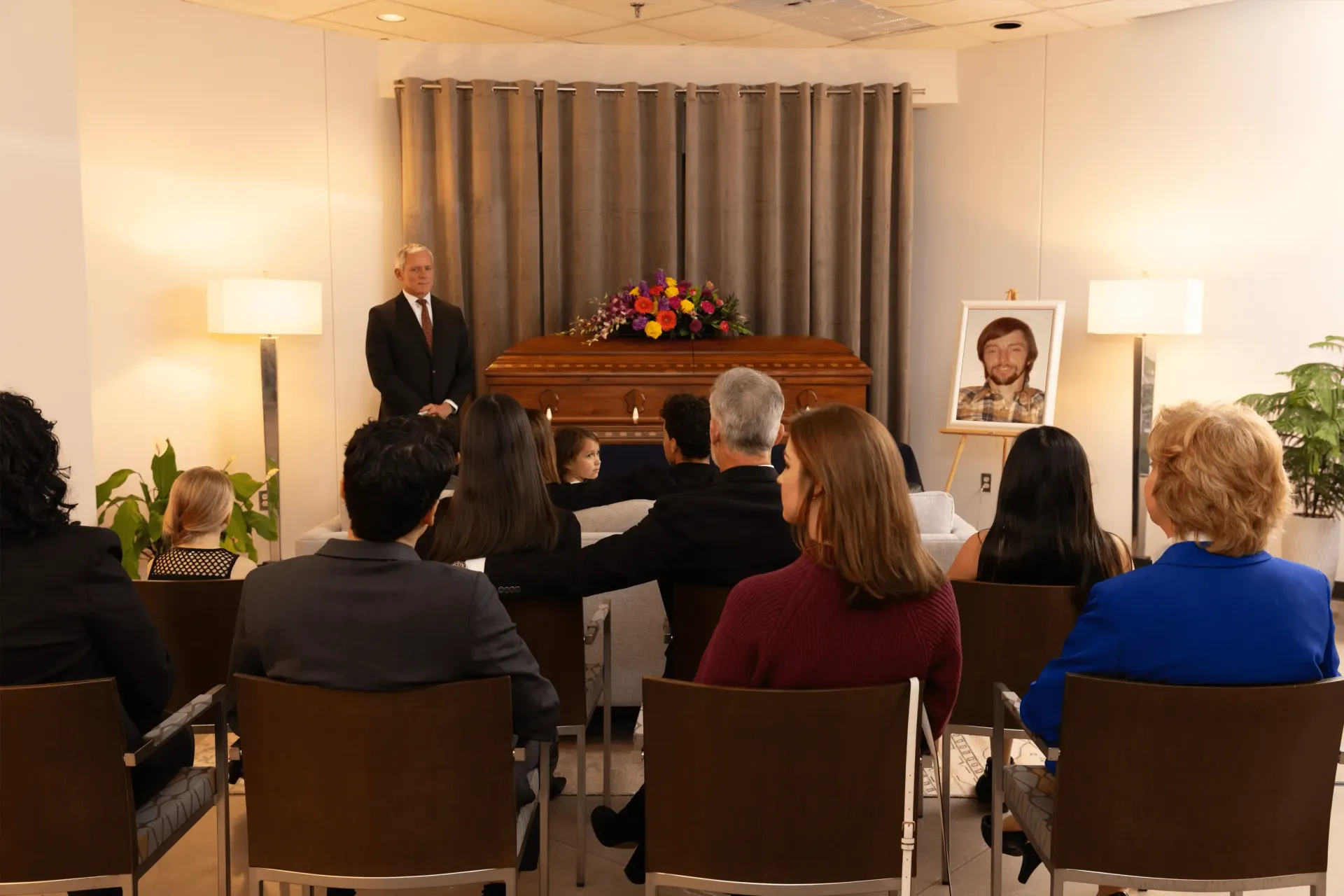 Mourners seated at a memorial service, facing a casket beside a framed portrait and floral arrangement.