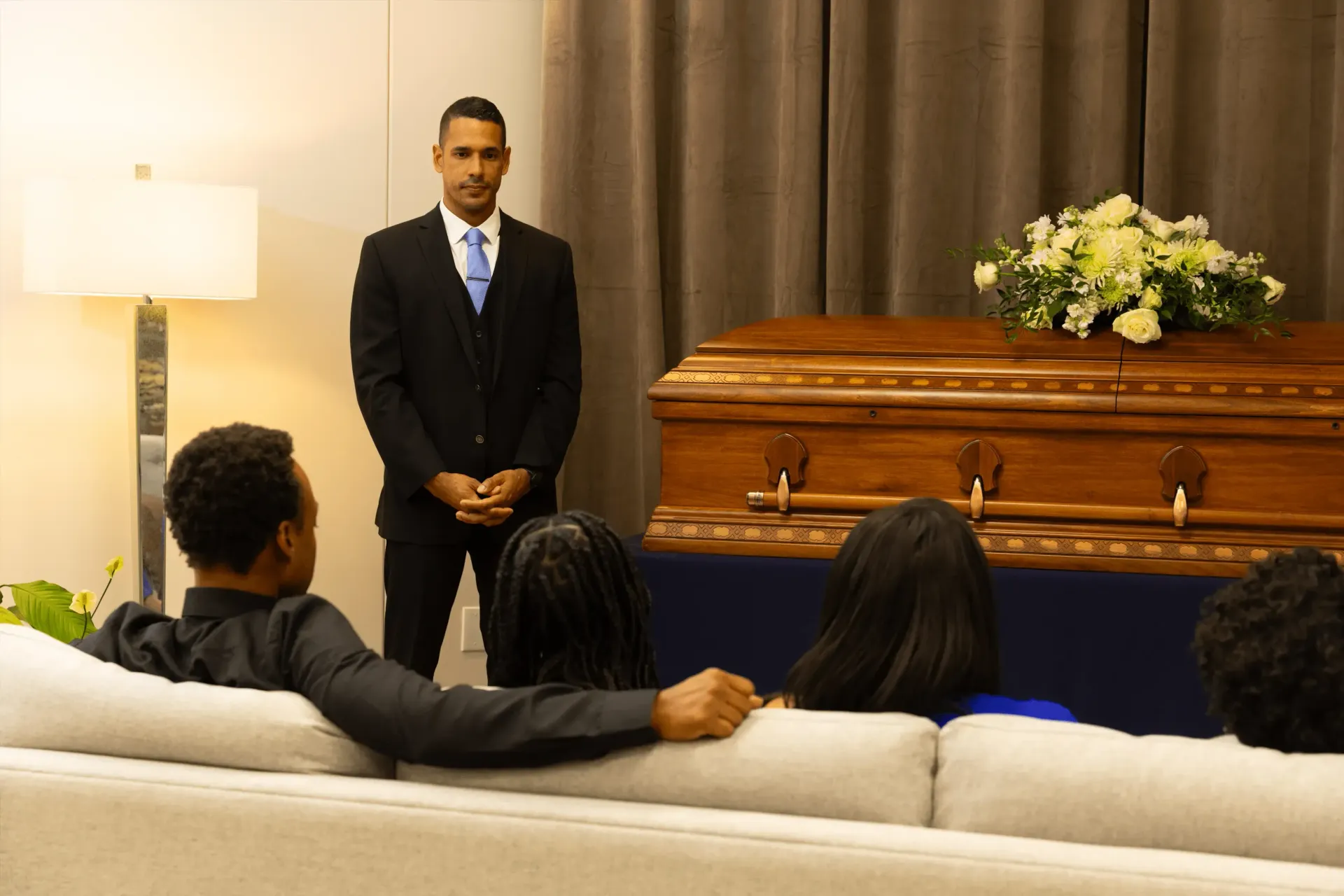 Mourners seated in a funeral home listening as a man speaks beside a casket adorned with flowers