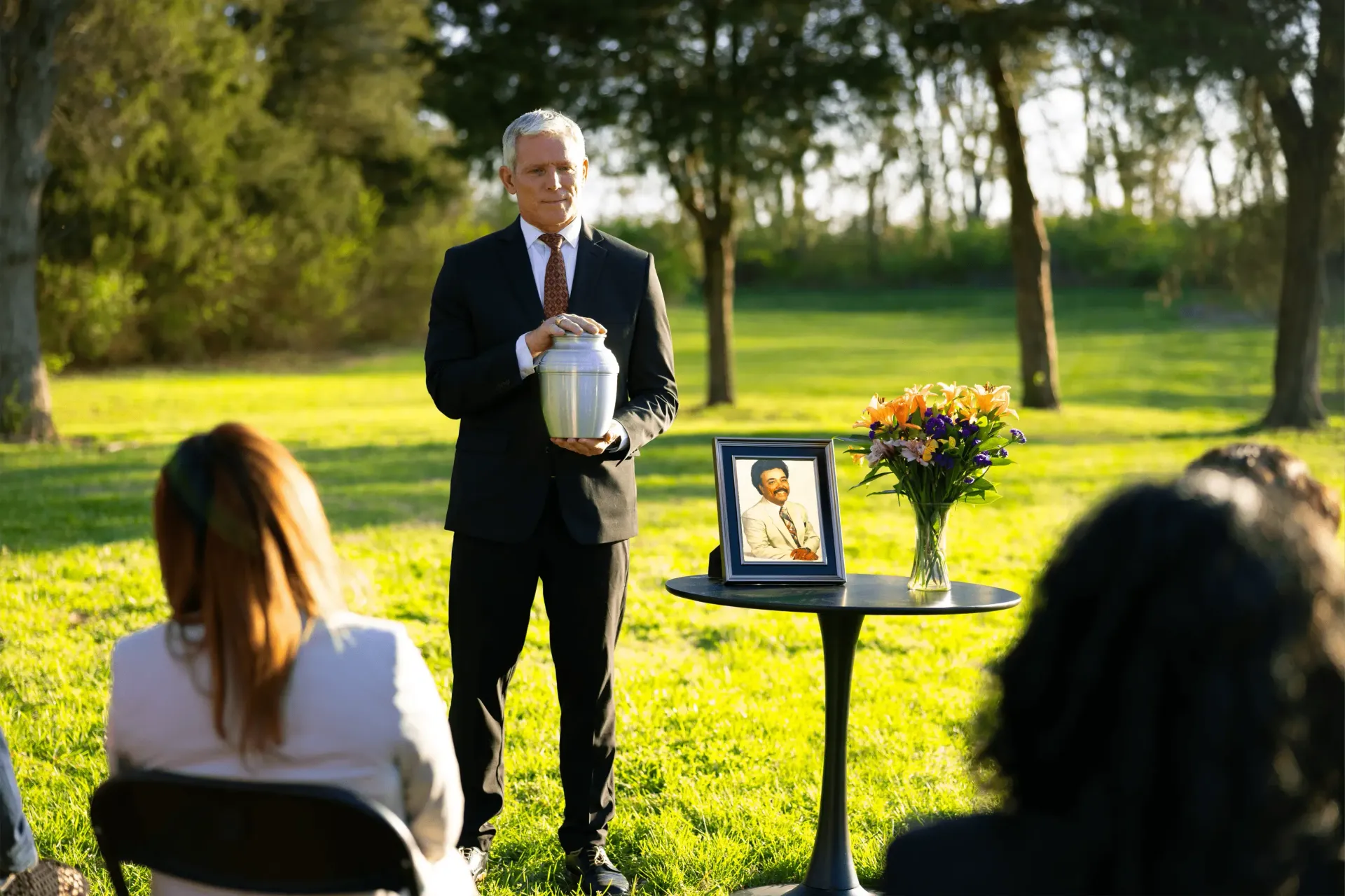 Outdoor memorial service with a man holding an urn beside a framed photo and flowers on a small table