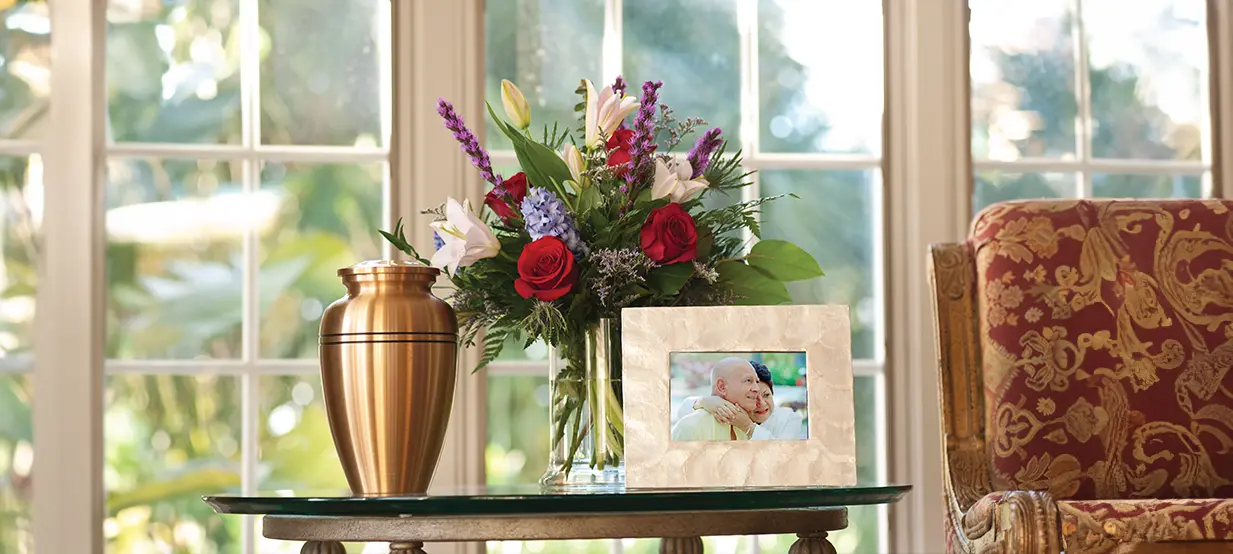 Vase of flowers and gold urn on a glass table by a sunlit window, with a patterned chair beside it