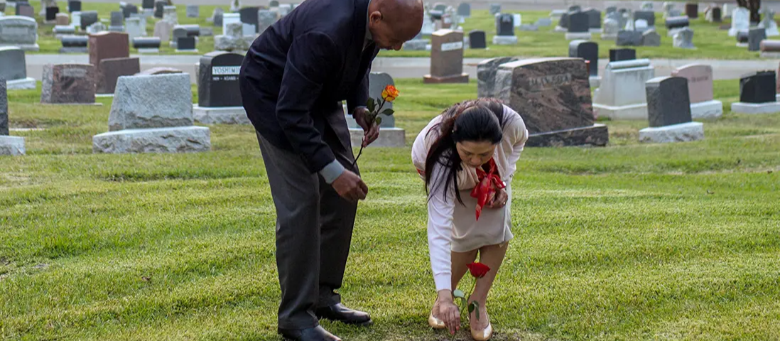 Two people kneel at a gravesite in a cemetery, placing flowers among headstones.