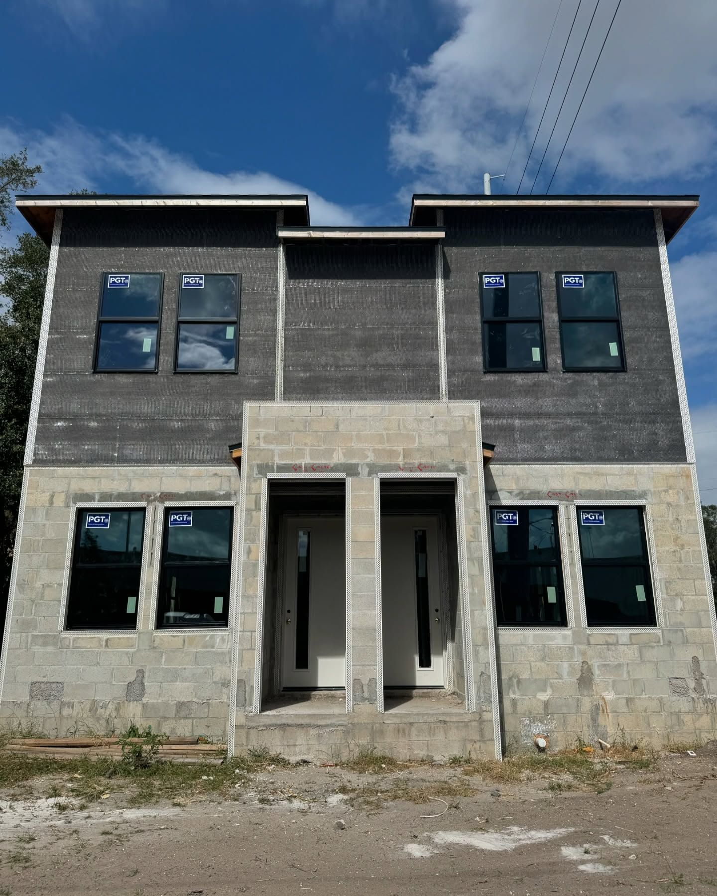 Two-story duplex under construction with cinder block base, black windows, and gray siding. Bright blue sky.