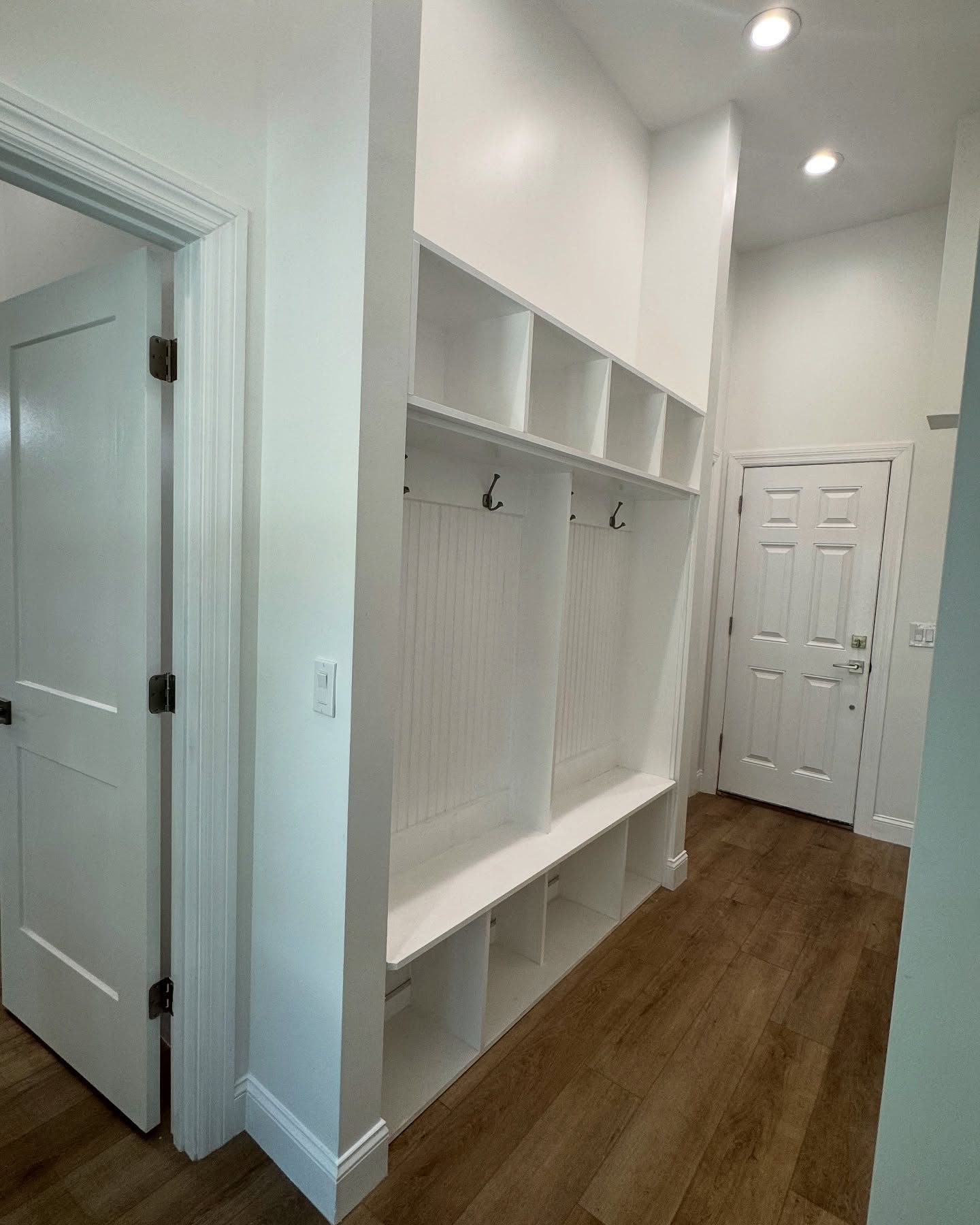 White hallway with built-in storage lockers, hooks, and bench, wooden floor. A door on each side.