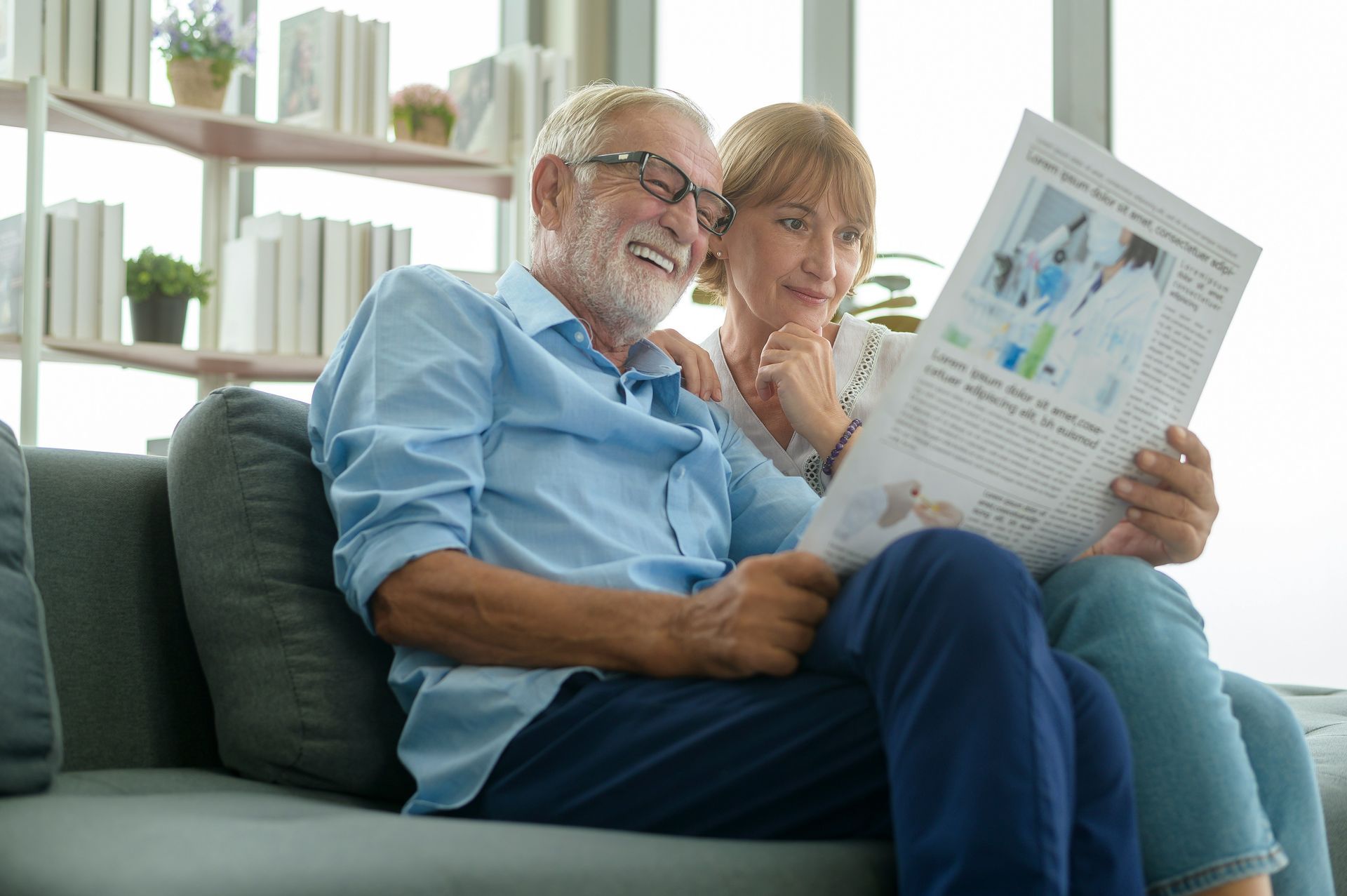 An elderly couple is sitting on a couch reading a newspaper.
