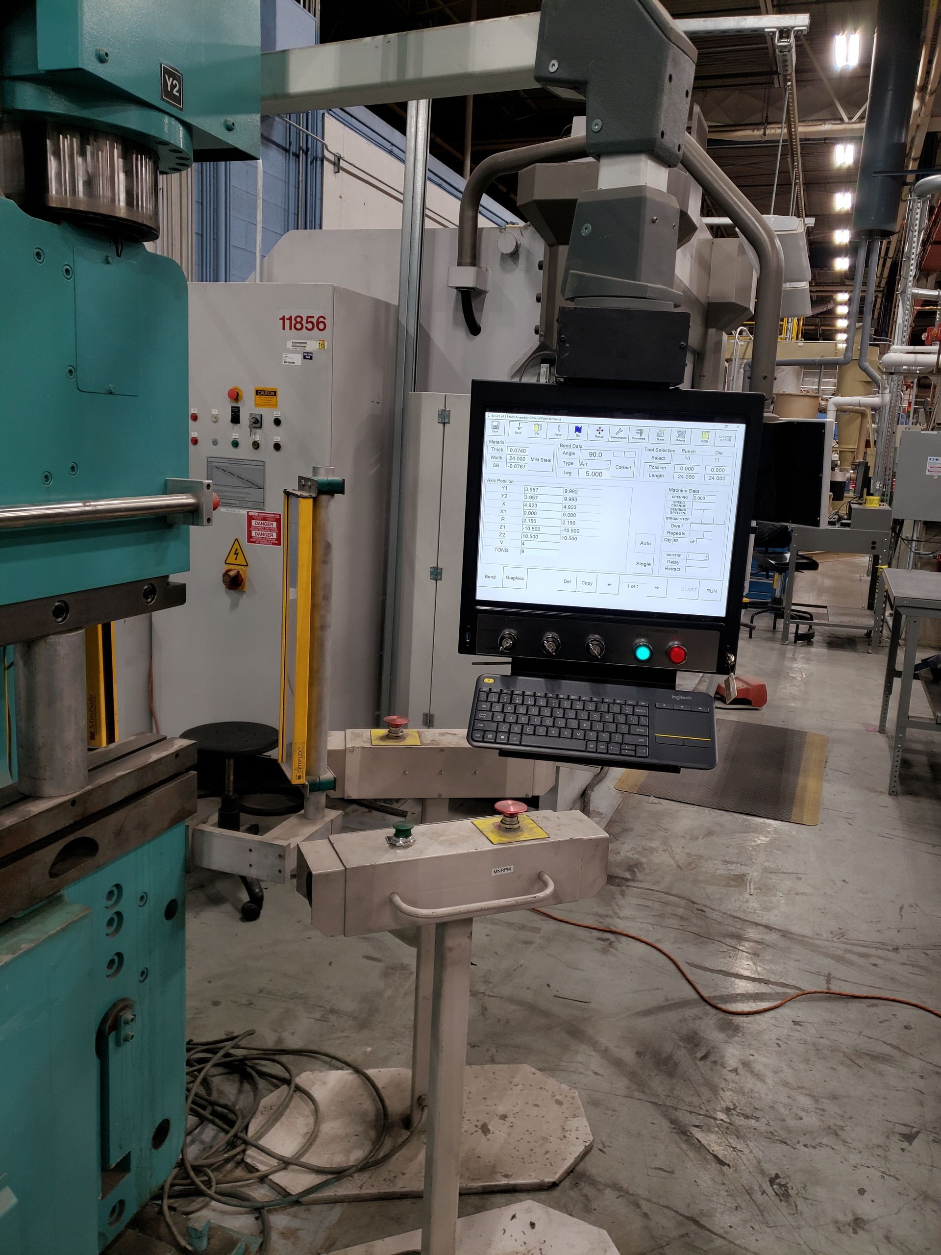 Industrial machine control panel with lit screen in a workshop, surrounded by cables and metal equipment.