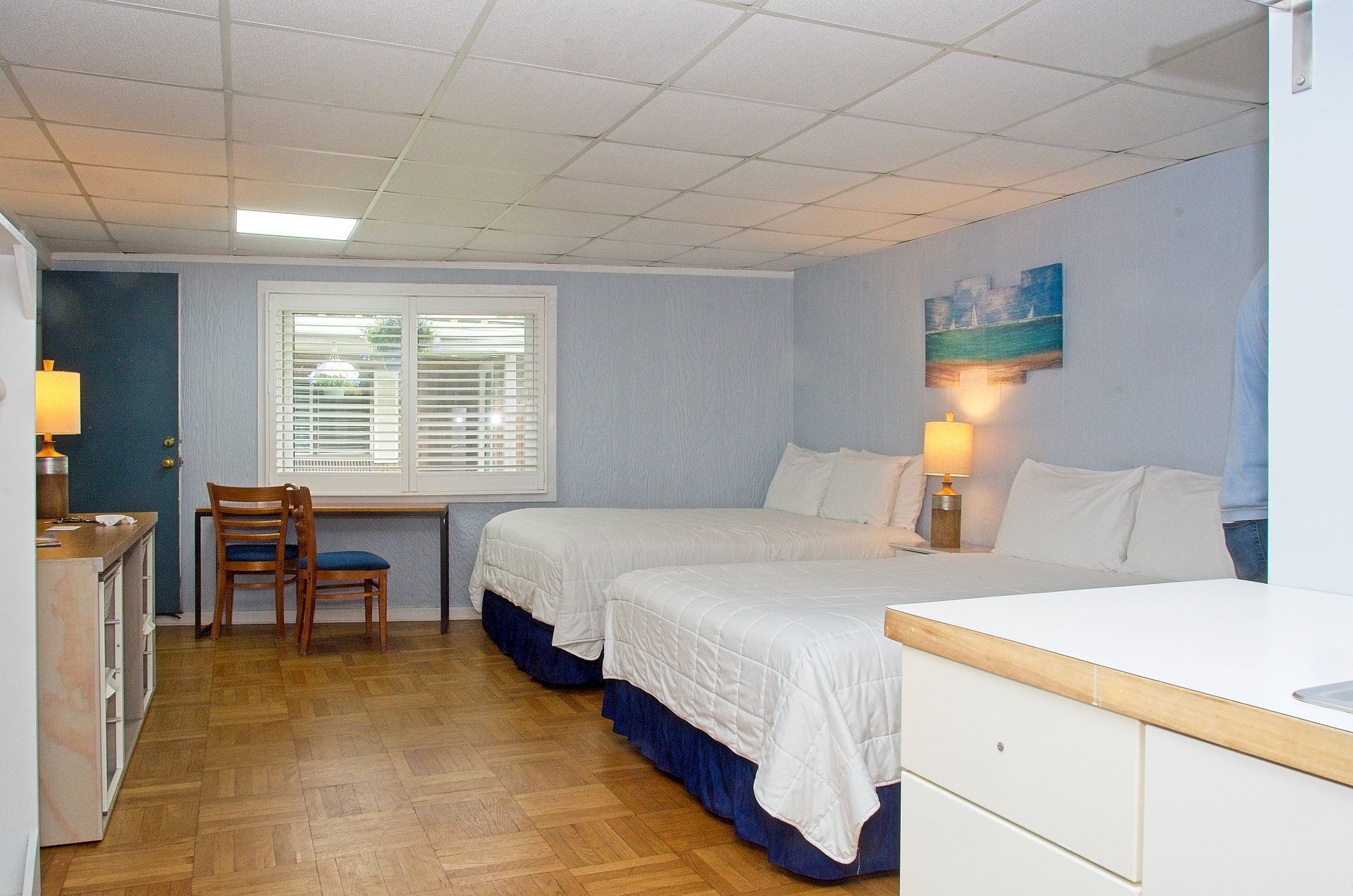 Bedroom with two double beds, light blue walls, a window, and a desk. There's a cabinet with a TV on the left side and light wood floors.