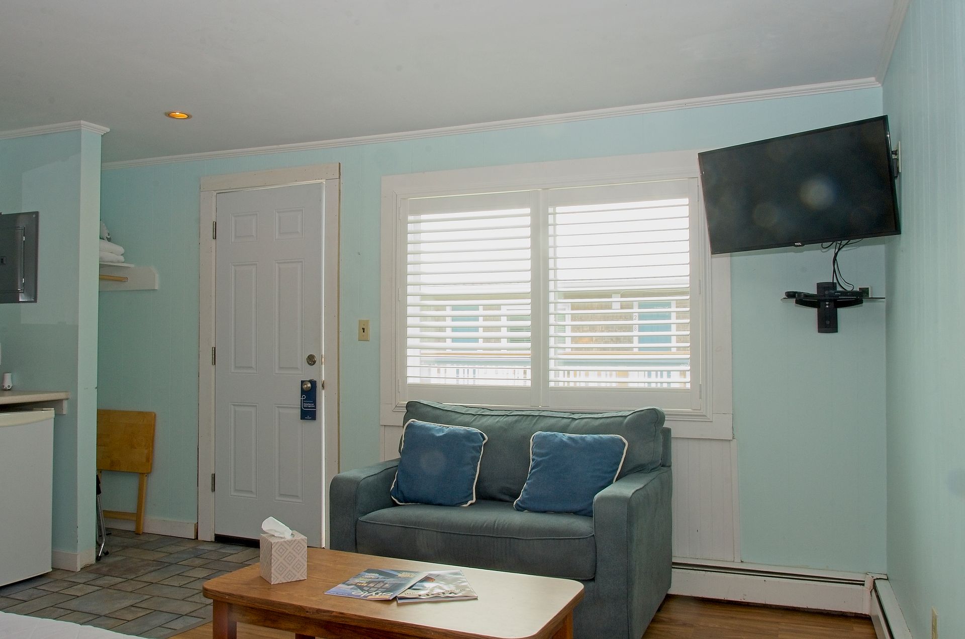 A motel room with a small couch, a window with shutters, and a mounted TV on a light blue wall.