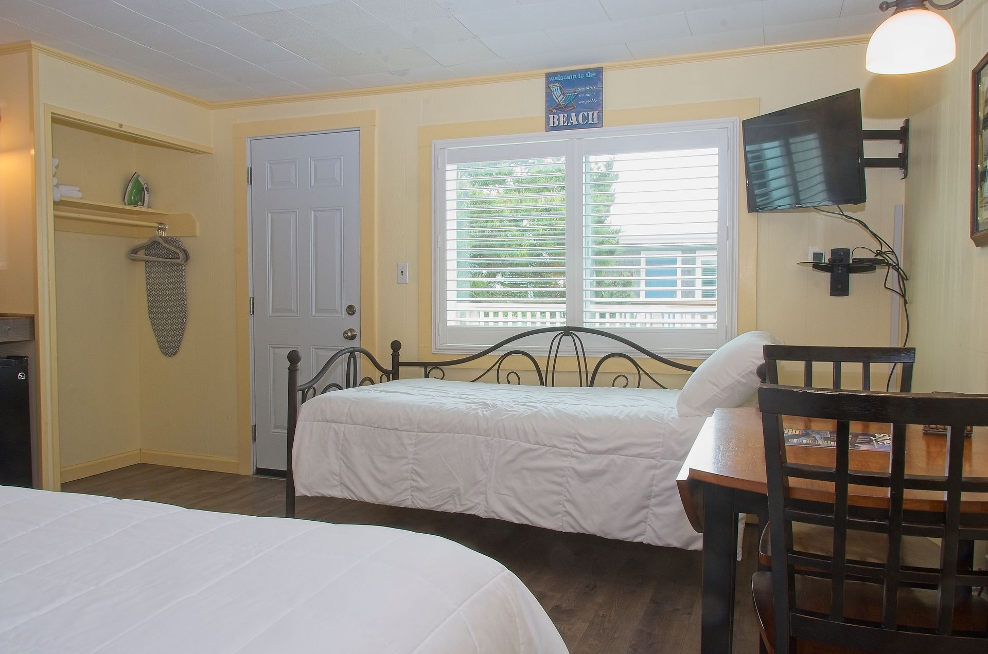 A small hotel room with a double bed, daybed, TV, and window overlooking trees. The walls are yellow and the bedding is white.