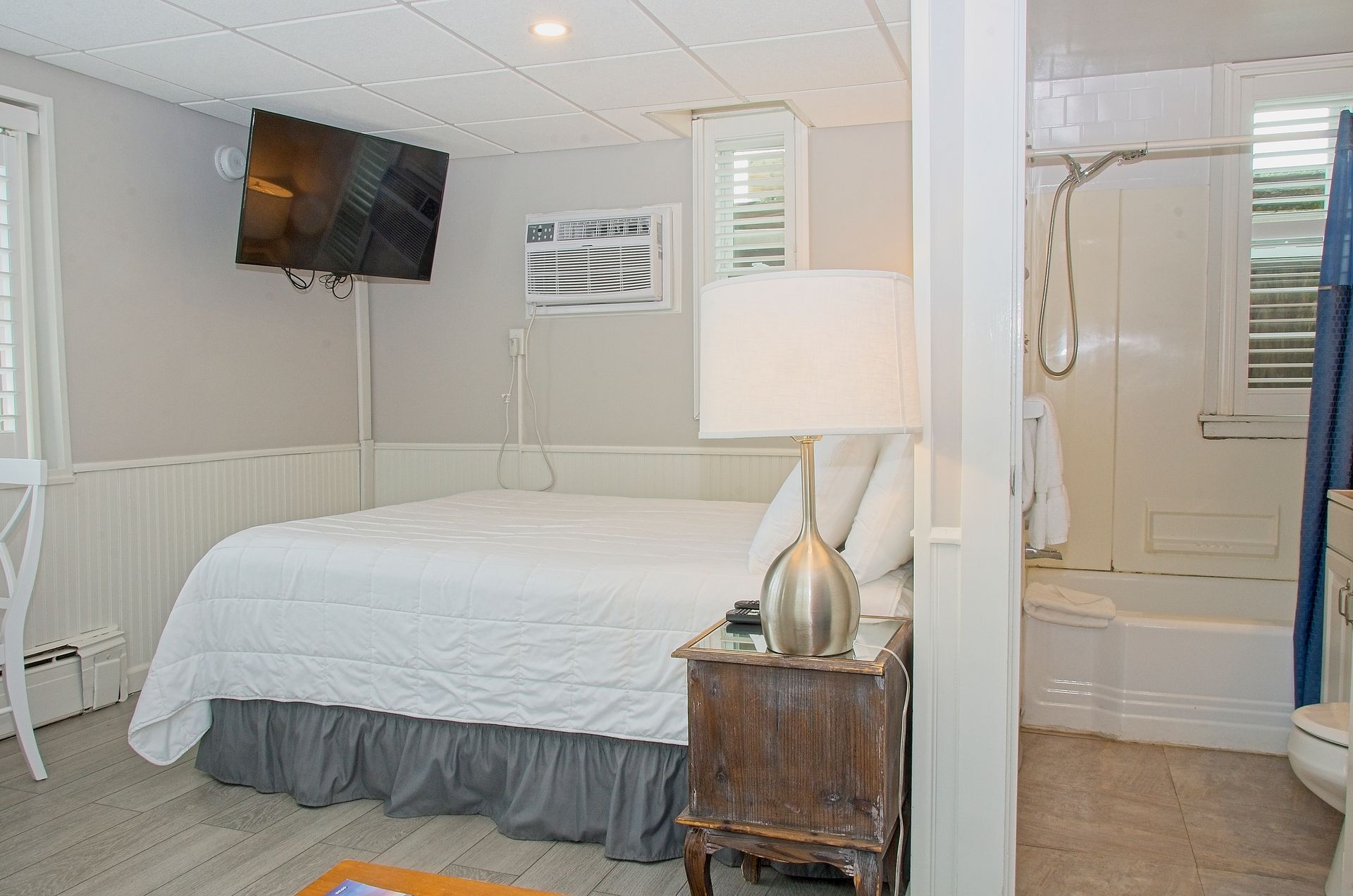 Bedroom with a bed, nightstand, television, and a bathroom visible to the right. The room is painted white and grey.