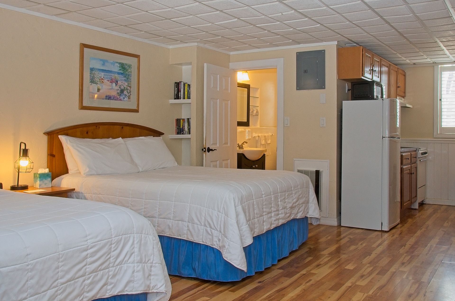 A small studio apartment with two beds, kitchenette, and a bathroom. The room has light-colored walls and wood flooring.