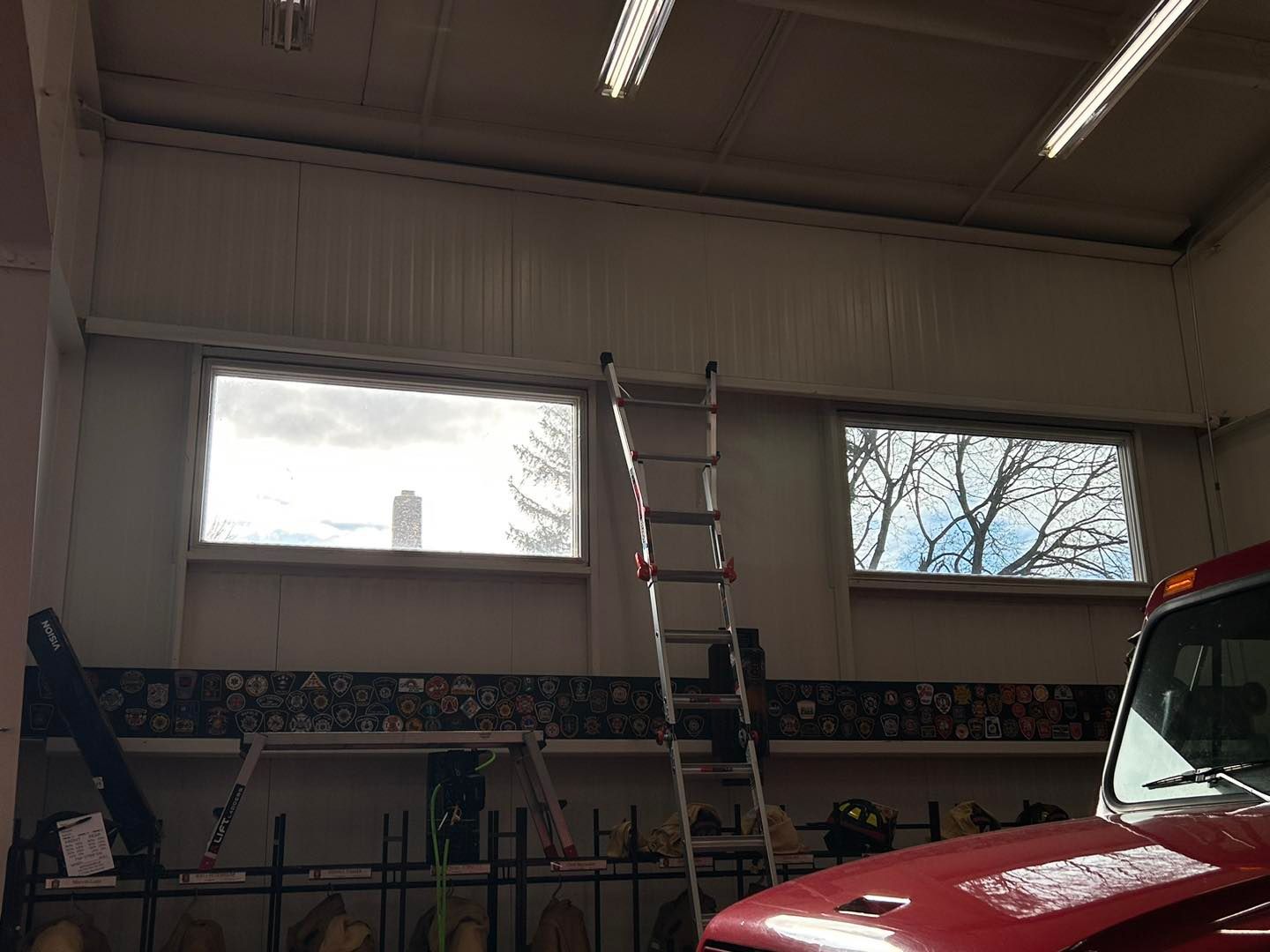 An extension ladder stands against an interior wall with two large windows in a room with a partial view of a red vehicle.