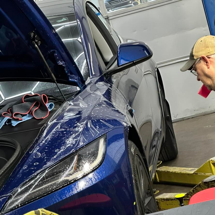 A worker applies a clear protective film to the fender of a dark blue car in a garage with the hood open.