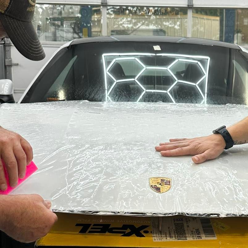 Two people apply clear protective film to the hood of a white Porsche in a workshop with geometric ceiling lighting.