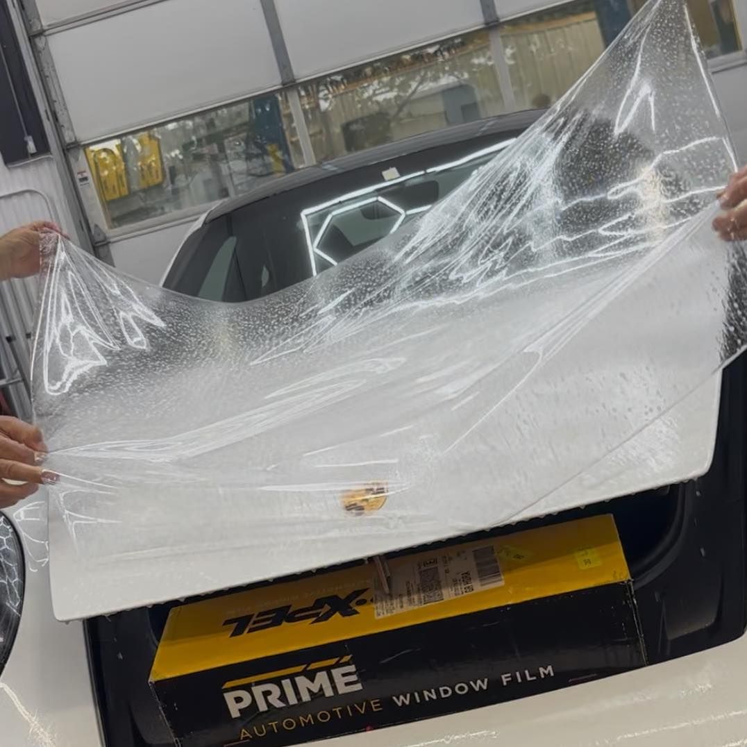 Two people hold a sheet of clear protective film over the trunk of a white car, with a box of window film in the trunk.
