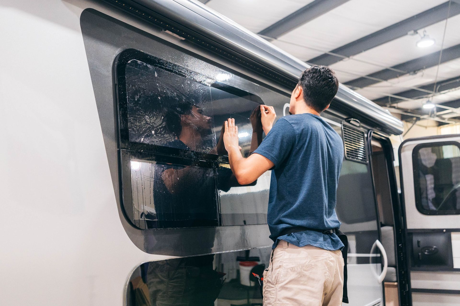 A person applying a window tint film to the side window of a recreational vehicle in a workshop.