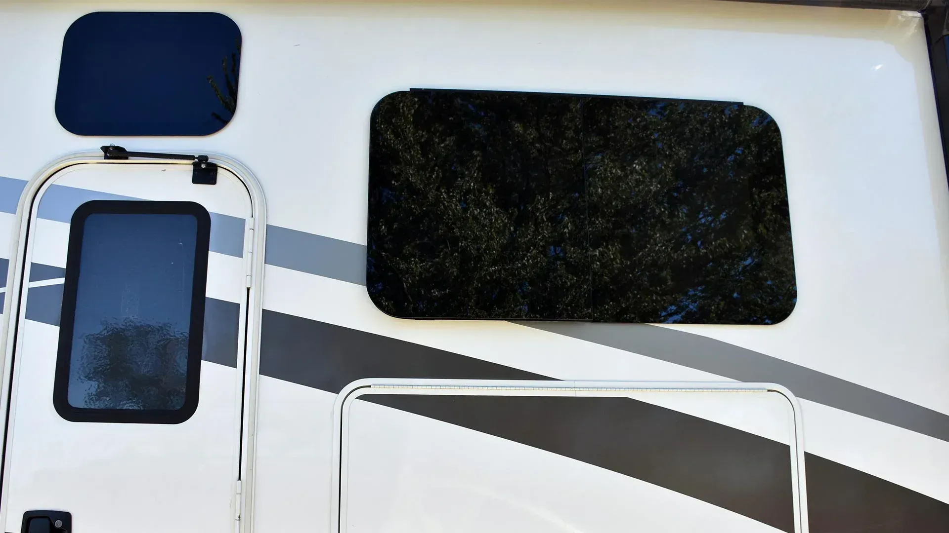 Exterior side view of a recreational vehicle with a door, a small window, and a larger tinted window over a grey graphic.